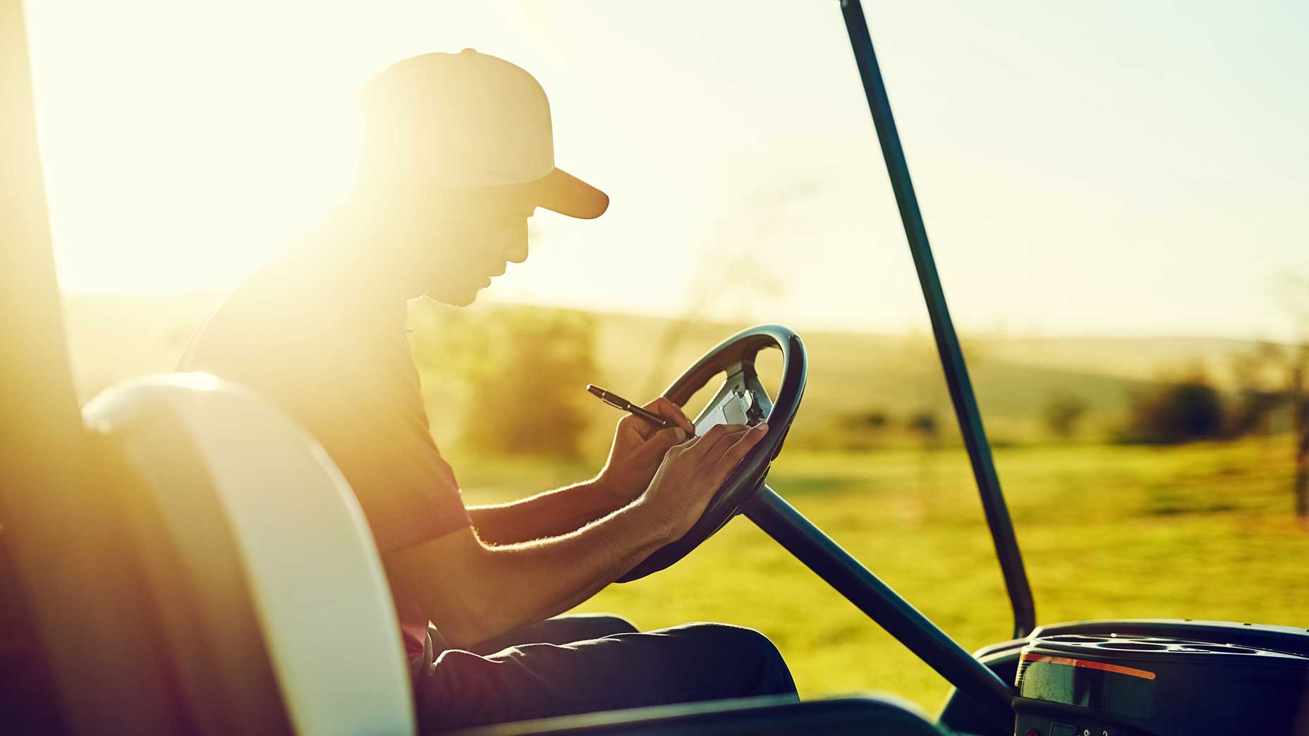 Golfer writes in scorecard while sitting in golf cart at sunset